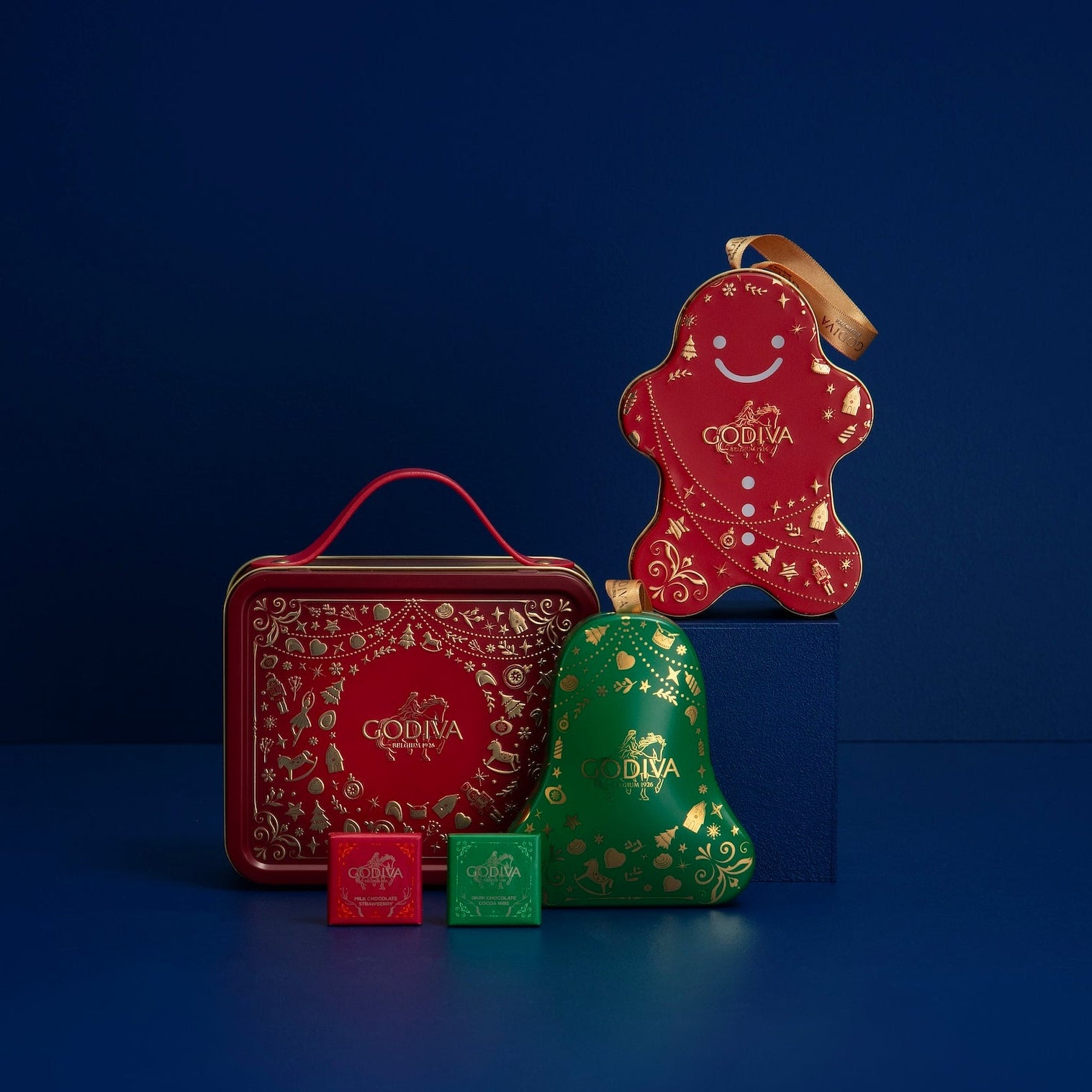 Godiva Christmas-themed chocolate packaging including a gingerbread man, bell, and tin on a blue background.